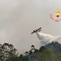 Aereo caccia in prova di fuoco sparando spruzzi d’acqua su pendio montuoso, nuvole grigie sul cielo.