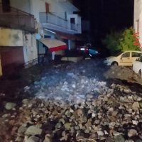Strada disastrata e pietrisco sparso in mezzo, edifici a lato, auto parcheggiate, scena notturna illuminata da luci occasionali