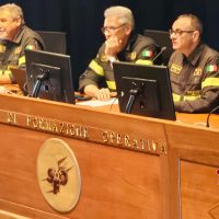 Tre pompieri in divisa, seduti dietro una scrivania, in conferenza, con microfoni e monitor, pronto a intervenire.