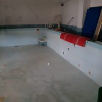 Interno di una piscina vuota con bordo bianco e sedili rossi, luci soffuse e pareti neutre, area di nuoto non in uso.