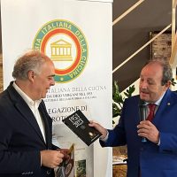 Due uomini in abiti scuri si scambiano una stretta di mano durante un evento; lo sfondo mostra banner con simboli ufficiali italiani e logo istituzionale.