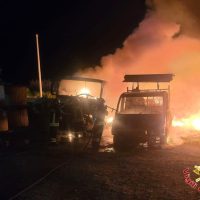 Automezzi ingorgati tra fiamme ardenti e fumo, scenario notturno, luci intense illuminano la zona, atmosfera di emergenza.