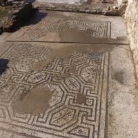Antico pavimento romano in pietra, motivi geometrici intricati e superfici levigate, immerso in luce solare.