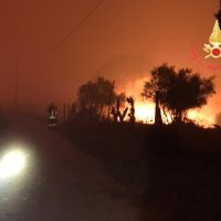 Incendio boschivo notturno, fiamme luminose tra gli alberi e una figura in divisa accanto a una strada.