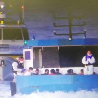 Gruppo di persone a bordo di un catamarano blu, al largo, con mare agitato e luci notturne sullo sfondo.