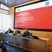 Tre relatori seduti a un tavolo di conferenza davanti a uno schermo con grafica Calabria 2032, palco e pubblico.
