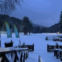 Scena invernale: piste innevate, alberi, forti e segnali verdi all’ingresso, luci serali oltre i palchi, atmosfera tranquilla e fredda.