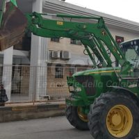 Trattore agricolo verde con caricatore frontale, operante in strada accanto a una casa, inquadratura ravvicinata su braccio meccanico.