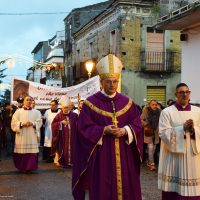 Predisposito sacerdote in veste viola dorata accompagna processione religiosa in una strada urbana con fedeli e architettura storica sullo sfondo.