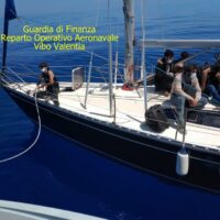 Velivolo di una barca a vela con persone a bordo, in mare azzurro; testo giallo in alto a sinistra: Guardia di Finanza Reparto Operativo Aeronavale Vibo Valentia.