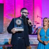 Uomo con barba e occhiali tiene microfono, sostiene trofeo circolare; donna in abito azzurro sorride accanto a lui in scena luminosa e colorata.