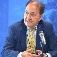 Uomo in giacca blu e cravatta gialla, parla al microfono durante una conferenza, sfondo blu sfumato.