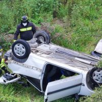 Incidente automobilistico: auto ribaltata su fianco in campagna con soccorritori in tuta gialla e nera che posizionano ruote sul mezzo.