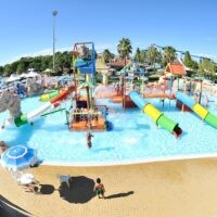 Parco acquatico con scivoli colorati, piscine azzurre e area sabbiosa; bambini giocano mentre genitori osservano in una giornata soleggiata.