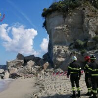 Due soccorritori in tuta antincendio osservano una frana su una spiaggia rocciosa, con cumuli di pietre e un cielo azzurro.