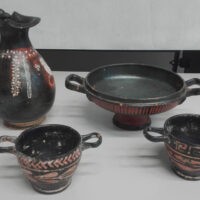 Set di ceramiche nere e rosse con decorazioni geometriche, tazza, piattino e tegame, disposti su piano bianco.