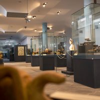 Interno di museo con teche e vetrine espositive illuminate, corridoi luminosi e visitatori che osservano reperti storici.