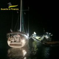 Immagine notturna: barca a vela ormeggiata con soccorritori della Guardia di Finanza vicino, luci sparse sull'acqua.