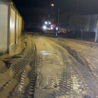 Strada sterrata notturna con pneumatici impronte, luci lontane e recinto a destra, atmosfera umida e silenziosa.