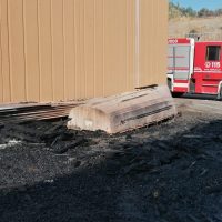 Cedimento del marciapiede accanto a una struttura gialla, con asfalto bruciato e veicolo di emergenza rosso sullo sfondo.