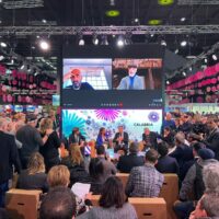 Conferenza affollata in sala grande: pubblico seduto, pannelli e relatori sul palco sotto schermi luminosi, atmosfera vivace e professionale.