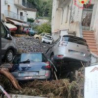 Crollo stradale in zona urbana: auto coinvolte tra detriti, strade danneggiate e edificio, persona curiosa in background.