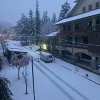 Strada innevata in un villaggio di montagna, auto parcheggiata, USBs di luci, edifici in legno, pini coperti di neve e cielo grigio.