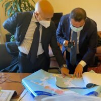 Due uomini in abiti formali esaminano mappe e documenti su un tavolo, con oggetti da ufficio e una cartina aperta.