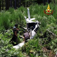 Incidente aereo in campagna: rottami della navicella personale tra erba alta, due persone vicine al relitto, bosco sullo sfondo.
