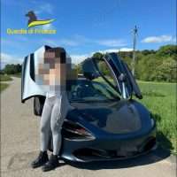 Auto sportiva nera su strada di campagna, versione coupé con apertura porte a farfalla e persona con volto pixellato accanto, logo Queste in primo piano.