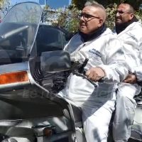 Due motociclisti seduti sul sidecar di una moto, indossano giacche chiare e occhiali, in un paesaggio urbano.