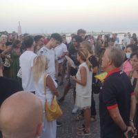 Gruppo di persone al mare durante un evento all'aperto, conversano tra di loro in abiti estivi in una giornata soleggiata.