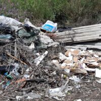 Rifiuti sparsi su un terreno con rami, legno e materiali di scarto, ambiente di discarica all'aperto.