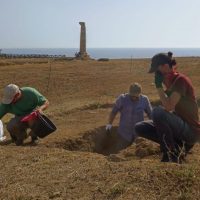 Gruppo di persone che scavano nel terreno davanti al mare, con un’antena o torretta in pietra sullo sfondo.