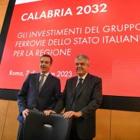 Due uomini in abiti formali sorridono in scena davanti a uno schermo rosso con testo italiano: Calabria 2032, investimenti Ferroviario di Stato.