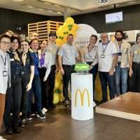 Gruppo di persone in abiti casual con mascotte e tavolo stile McDonald's, brindano attorno a una torta in uno spazio interno.