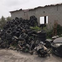 Scatolone di pneumatici usati accatastati accanto a una struttura in rovina, con motori e macchinari sommersi dalla vegetazione.