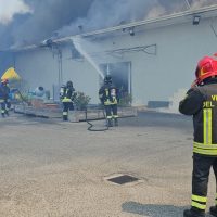 Vigili del fuoco in azione mentre spegnono un incendio in un edificio industriale, con fumo visibile e personale coordinato sul posto.