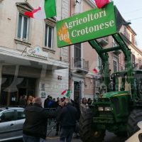 Gruppo di agricoltori con trattore verde e cartelli 'agricoltori del Crotonese' durante una manifestazione in strada, vicino a edifici storici.