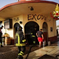 Due vigili del fuoco all’ingresso di un edificio rotondo in stile messicano, con graffiti e persone sul portico, intervento d’emergenza.
