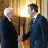 Due uomini di fronte e strettamente stretti la mano in un corridoio dorato, atmosfera formale e calorosa.