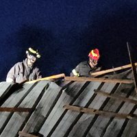 Due operai in caschi lavorano su un tetto inclinato in legno, impugnando attrezzi durante il lavoro notturno.