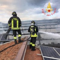 Due vigili del fuoco indossano tute riflettenti su tetto con pannelli solari, fronteggiando un incendio sullo sfondo.