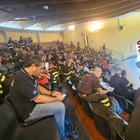 Sala affollata di spettatori seduti, molte persone con magliette scure e una scena illuminata sul palco.