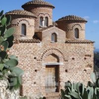 Chiesa in stile bizantino in pietra, cupole rotonde, contesto siciliano con cactus e paesaggio collinare sullo sfondo.