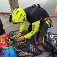Tecnico alpinista in casco giallo prepara corde e accessori su una parete rocciosa, attrezzatura di sicurezza pronta per l’arrampicata.