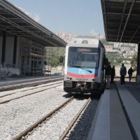 Treno pendolare su binari con piattaforma e due persone in attesa, stazione situata in una zona collinare.