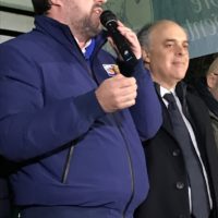 Uomini in abiti formali e pubblico, uno parla al microfono durante un evento pubblico.