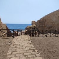 Sentiero di pietra che conduce al mare tra vecchie mura di castello, con ponte e barriere in legno, cielo azzurro