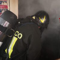 Pompiere in tuta nera, estintore e attrezzature, affronta fumo denso in ambiente sospeso, luce gialla sul casco.